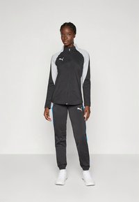 Fekete és szürke sportos melegítőszett, cipzáras dzsekivel és szűkített szárú nadrággal, kék részletekkel és Puma logóval. Könnyű anyagból készült.