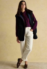 Kobieta stojąca przy beżowej ścianie, ubrana w bordową koszulę w wzory, czarną bezrękawnik, białe spodnie i brązowe mokasyny.