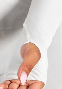 Close-up van een hand met roze nagellak door de duimopening van een witte lange mouw shirtmouw met geribde manchet.