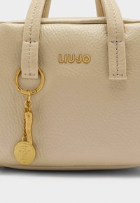 Beige Lederhandtasche mit goldenen Beschlägen, ausgestattet mit geprägtem Logo, zwei Griffen und einem dekorativen Anhänger, der an einem Metallring befestigt ist.