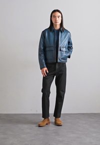 Chaqueta de cuero azul con dos bolsillos frontales, combinada con pantalones negros y zapatos marrón claro. Textura suave, diseño estructurado y líneas limpias.