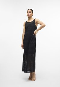 Vestido longo preto sem mangas possui uma textura de renda com padrões horizontais. Está estilizado com saltos em tom nude e acessórios.
