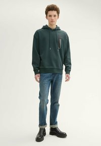Ciemnozielona bluza z kapturem, z trokami, kieszenią z przodu i graficznym wzorem. Połączona z niebieskimi dżinsami i czarnymi botkami do kostek.