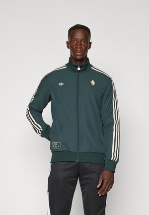 Veste de survêtement zippée verte avec design à trois rayures blanches et logo de lion, col et poignets côtelés, tissu doux, poches avant et étiquette de marque.