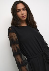 Schwarzes Kleid mit kurzen Ärmeln, bestehend aus solidem Stoff und durchscheinender, bestickter Spitze. Es hat eine taillierte Taille und eine entspannte Silhouette.