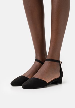 Jambes portant des chaussures à talons bas en suede noir avec une bride à la cheville et des pointes contre un fond blanc.