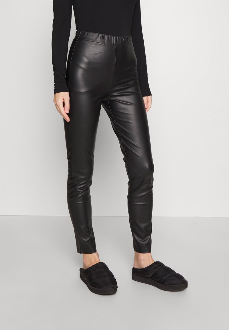 Black faux leather leggings with a high elastic waistband, fitted silhouette, and smooth texture. Paired with black quilted slip-on shoes.