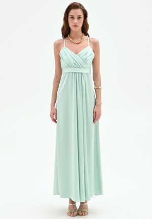 Giovane donna che indossa un vestito verde menta lungo fino a terra senza maniche con corsetto arricciato, collana d’oro, braccialetto e sandali, in piedi davanti a sfondo bianco.