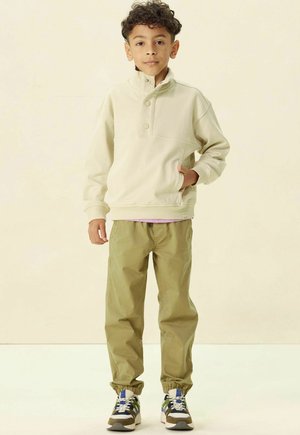 Helles beige Pullover mit Stehkragen und zwei Knöpfen, kombiniert mit olivgrünen elastischen Bündchenhosen und mehrfarbigen Sneakers.