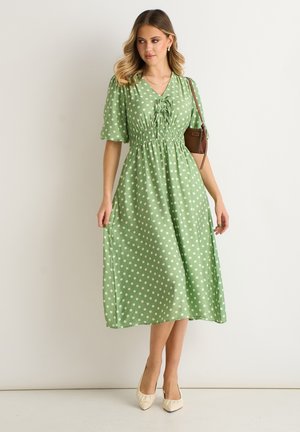 Woman wearing a green midi dress with white polka dots, puff sleeves, and a gathered waist, carrying a brown shoulder bag and beige heels.
