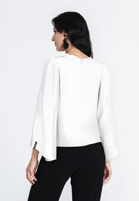 Weiße Bluse mit puffierten Ärmeln, glatter Textur und einem Reißverschluss hinten, kombiniert mit schwarzen Hosen. Einfaches, modernes Design.