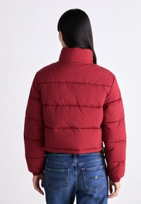 Röd puffjacka med hög krage, med horisontella kviltade sektioner och elastiska manschetter. Matchas med blå denimjeans.
