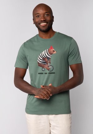 T-shirt en coton vert avec un graphique d'un ours portant une chemise rayée et un chapeau rouge, chevauchant un vélo, avec du texte en dessous en noir.