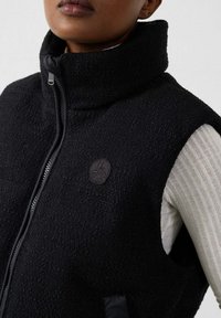 Primo piano di una persona che indossa un gilet nero con cerniera e trama, sopra una maglia bianca a costine con maniche lunghe, che mostra il colletto alto del gilet e la toppa con il logo.
