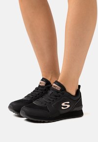 Person som bär svarta Skechers-sneakers med rosa logotyp och detaljer, stående på en ljus bakgrund med bar ben synliga.