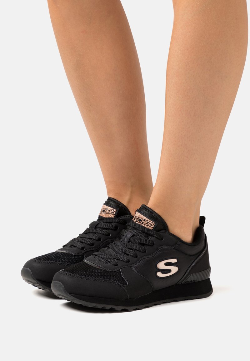 Person som bär svarta Skechers-sneakers med rosa logotyp och detaljer, stående på en ljus bakgrund med bar ben synliga.