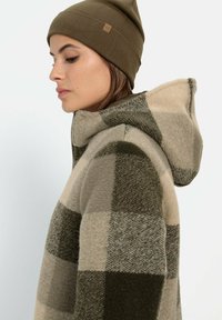 Karierte Fleecejacke mit Kapuze in Beige und Olivgrün, mit einem Reißverschluss. Getragen mit einer passenden Mütze in Olivgrün.