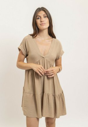 Jeune femme aux longs cheveux bruns portant une robe ample beige à manches courtes avec un décolleté en V profond, debout devant un fond clair uni.
