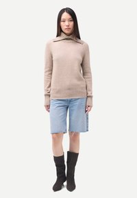 Femme aux longs cheveux bruns portant un pull à col roulé beige, un short en denim bleu clair et des bottes noires mi-mollet, debout sur un fond blanc.