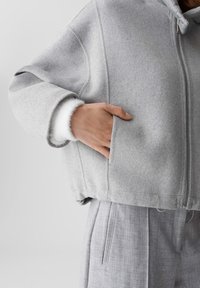 Hand in der Tasche einer hellgrauen Zip-Jacke mit weißem, flauschigem Bündchen, kombiniert mit hochtaillierten grauen, gefalteten Hosen.