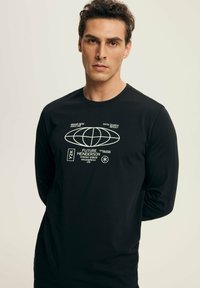 Man wearing black long-sleeve shirt with white futuristic graphic and text, standing against plain light background, hands behind back.