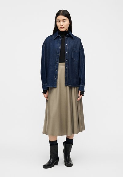 Γυναίκα που φοράει σκούρο τζιν πουκάμισο με κουμπιά πάνω από μαύρο ζιβάγκο, μπεζ μίντι φούστα, σκούρες κάλτσες και μαύρες μποτάκια μέχρι τη μέση της γάμπας, στέκεται μπροστά σε λευκό φόντο.