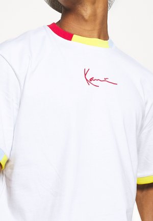 Witte t-shirt met meerkleurige kraag en mouwen in rood, geel en lichtblauw. Heeft een rode geborduurde handtekening op de borst.