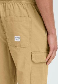 Khaki cargo nohavice s elastickým pásom, bočnými a zadnými vreckami a vyšitým štítkom. Textúrovaná bavlnená látka, neformálny dizajn.