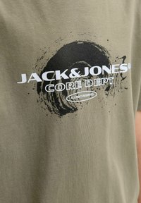 JACK & JONES CORE DEPT SVA PRAVA ZADRŽANA u bijeloj boji preko crnog kružnog poteza četkicom na maslinasto zelenoj tkanini.