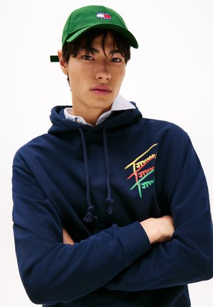 Sweat à capuche bleu marine avec un logo multicolore et un cordon de serrage. Associé à une casquette verte avec un patch logo. Le mannequin se tient les bras croisés.