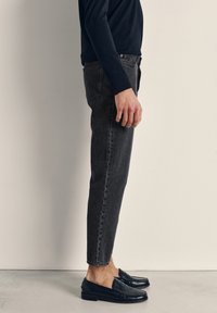 Moški stoji v pristriženi črni jeansi, črnih loaferjih in dolgi črni srajci z dolgimi rokavi na enemostenem svetlem ozadju.