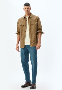 Camisa marrón de botones con dos bolsillos en el pecho, camiseta beige debajo, jeans azules y botas de cuero marrón. Modelo de pie contra un fondo blanco.