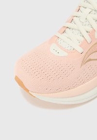 Zapatilla deportiva de color rosa claro con parte superior de malla, cordones planos y una puntera texturada. Cuenta con una suela de goma y acentos de marca sutiles.