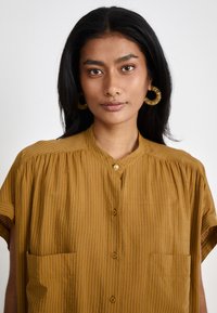 Camicia a maniche corte di colore senape con righe verticali, chiusura con bottoni sul davanti e due tasche sul petto. Tessuto leggero e strutturato.