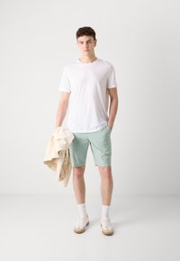 Hvid t-shirt, mintgrønne shorts og hvide sneakers med beige detaljer. Modellen holder en beige jakke; står foran en ensfarvet baggrund.
