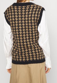 Pletený svetr bez rukávů v černobílém houndstooth vzoru, texturovaná látka, kulatý výstřih, nošený přes bílou košili.