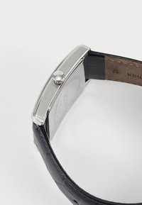 Boîtier de montre en métal argenté rectangulaire avec un bracelet en cuir noir texturé. Présente une couronne sur le côté et une finition polie.