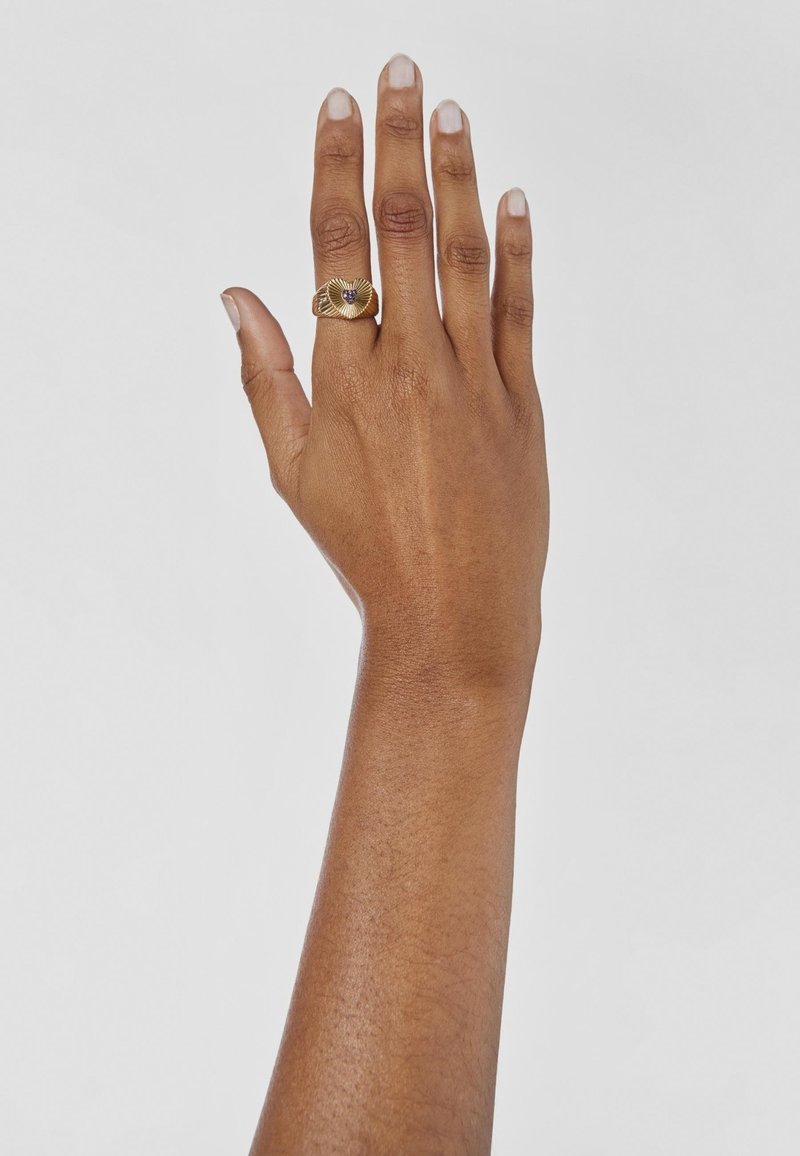 Anello d'oro con un design testurizzato e ondulato che presenta un gemma centrale, indossato su una mano marrone su uno sfondo bianco.