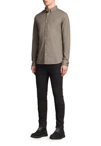 Mężczyzna ubrany w beżową koszulę z guzikami, czarne slim-fit spodnie oraz czarne buty na sznurowadła, stojący z rękami swobodnie opuszczonymi wzdłuż ciała.