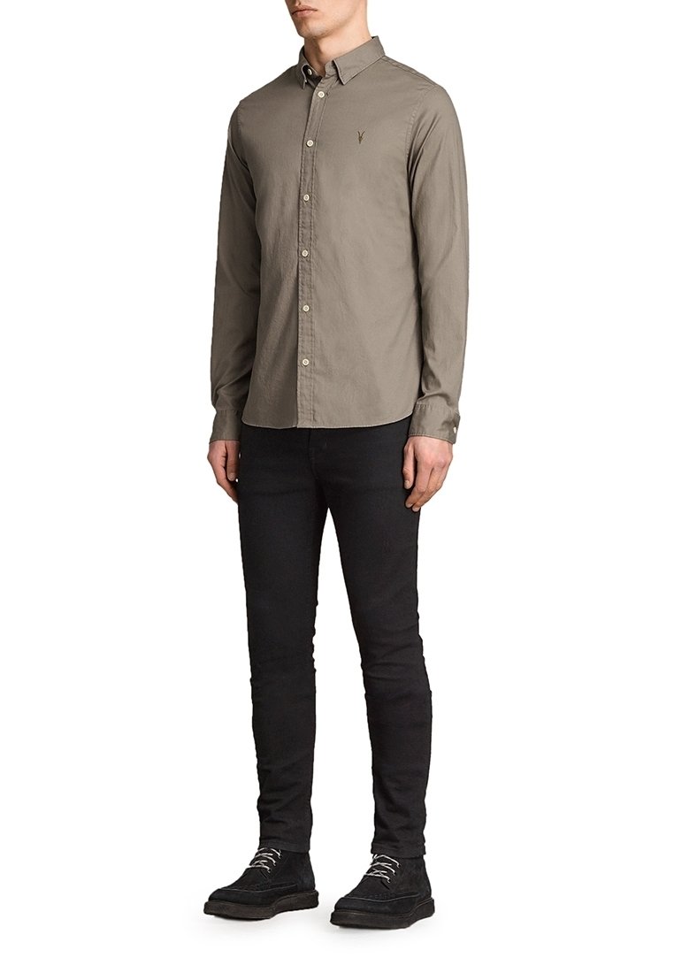 Mężczyzna ubrany w beżową koszulę z guzikami, czarne slim-fit spodnie oraz czarne buty na sznurowadła, stojący z rękami swobodnie opuszczonymi wzdłuż ciała.