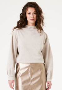 Sudadera beige con cuello alto, mangas largas y un detalle de cremallera lateral, combinada con una falda lápiz metálica dorada. Textura de tela suave.