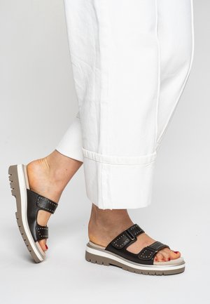 Schwarze Ledersandalen zum Herschieben mit zwei Schnallriemen, mit Nieten besetzt und mit einer dicken weißen und braunen Sohle. Getragen zu weiten weißen Hosen.