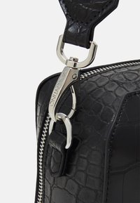 Detail černé texturované kožené tašky s kovovým zipem a klipem ve stříbrné barvě, doplněné hladkým popruhem.