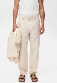 Beige Leinenhose mit lockerer Passform, ausgestattet mit einem Knopfverschluss und Seitentaschen, gehalten neben einem passenden Hemd. Braune Sandalen getragen.