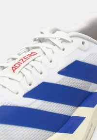 Baskets de course Adidas blanches avec des rayures bleues, des lacets blancs et une étiquette rouge "ADIZERO" sur la languette.