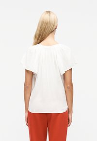 Γυναίκα με μακριά ξανθά μαλλιά που φοράει χαλαρό λευκό μπλουζάκι με κοντά παγωμένα μανίκια και κόκκινο παντελόνι, από πίσω, με φόντο λευκό.