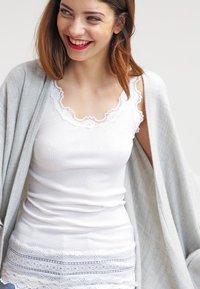 White ribbed tank top with lace trim at the neckline and hem, paired with a soft gray shawl, featuring an open front design.