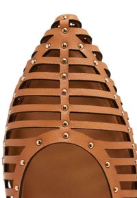 Zapato de cuero marrón con diseño ranurado y tachuelas doradas, que presenta un dedo del pie y un talón abiertos, enfatizando un patrón geométrico.