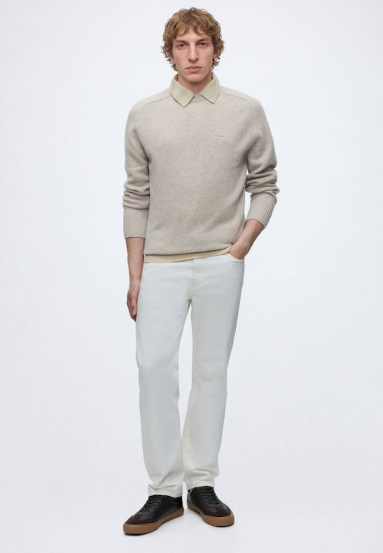 Homme aux cheveux bouclés debout portant un pull beige sur une chemise à col, un pantalon blanc et des baskets noires avec des semelles marron.
