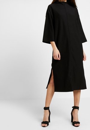 Vrouw draagt een zwarte midi-jurk met ellebooglange mouwen en een zijsplit, gecombineerd met zwarte hoge hak sandalen met enkelriem.